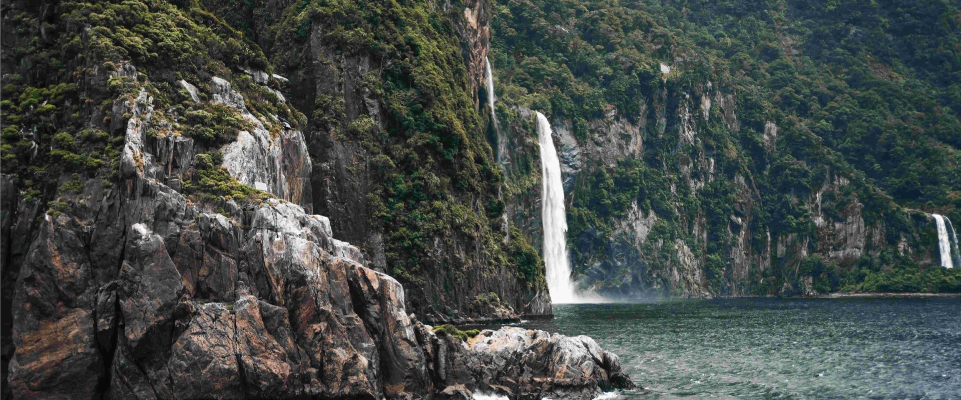 Waterfalls cascade into the fiord from cliffs green with trees and lush vegetation